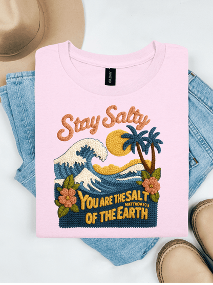 Pink t-shirt with embroidered design and text, placed on denim jeans with a beige hat and shoes in the background.