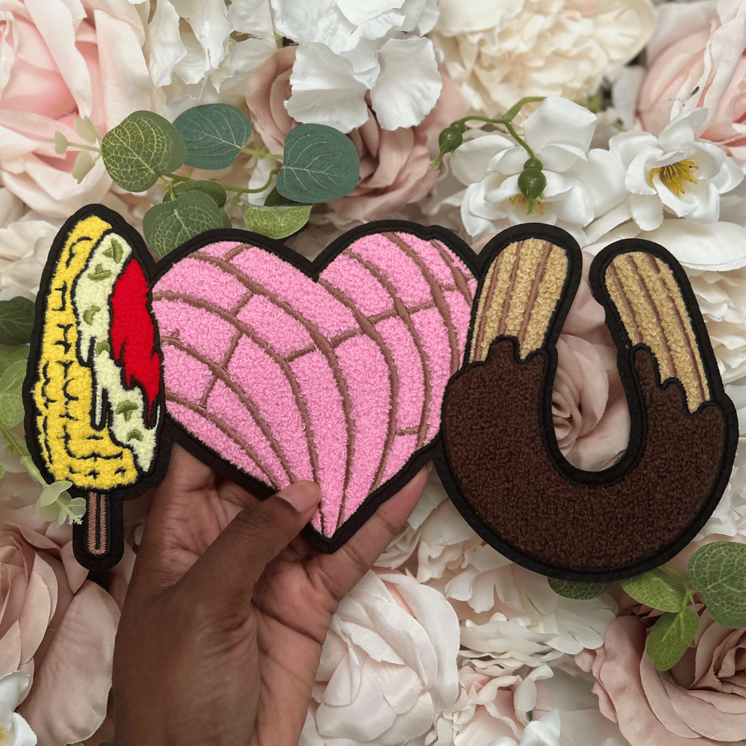 Three colorful patches shaped like a corn, pink heart, and brown donut held over a floral background.